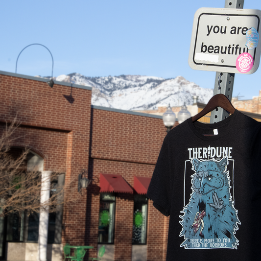 Black shirt with a graphic of a raccoon with an injured, bandaged arm. Text that says "There is more to you than the horrors" Hanging from a street sign that says "You are beautiful".