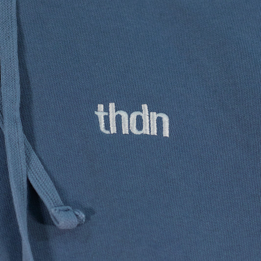 Close-up of a blue jean colored hooded sweatshirt laid flat. A white "thdn" logo is embroidered on the fabric.