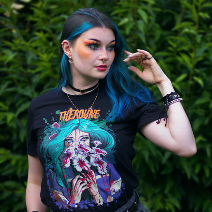 Black shirt with a stylized illustration of a person with teal hair and colorful makeup, worn by a model in front of bushes.