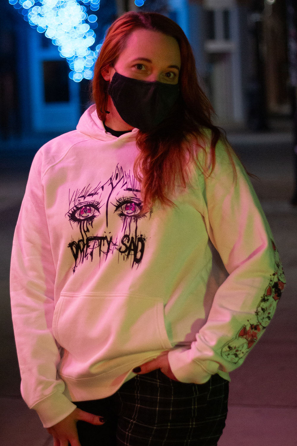 A white hoodie with a graphic print of stylized eyes and the text 'Pretty & Sad' on the front. Worn by a hoodie in an urban environment.