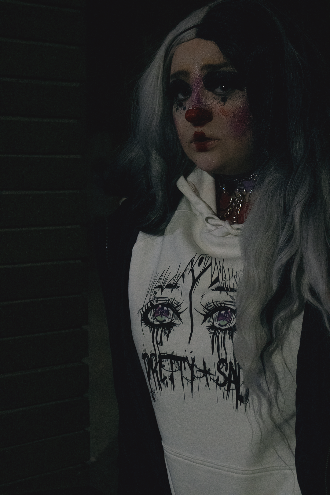 A person in clown makeup wearing a white hoodie with a graphic print of stylized eyes and the text 'Pretty & Sad' on the front.