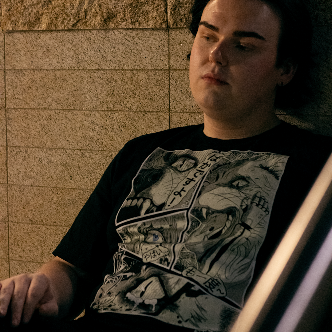 Black shirt with with 4 panel manga style illustration with japanese text and Therdune logo, worn by model, sitting against a brick wall.