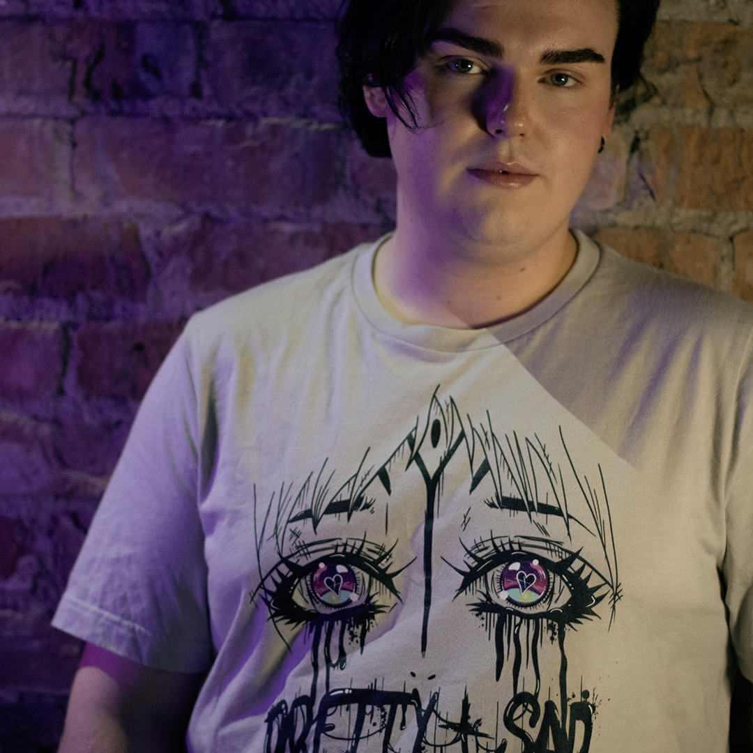 Cream color shirt with a graphic print of stylized eyes and the text 'Pretty & Sad' on the front. Worn by a model in front of a brick wall.