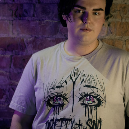 Cream color shirt with a graphic print of stylized eyes and the text 'Pretty & Sad' on the front. Worn by a model in front of a brick wall.