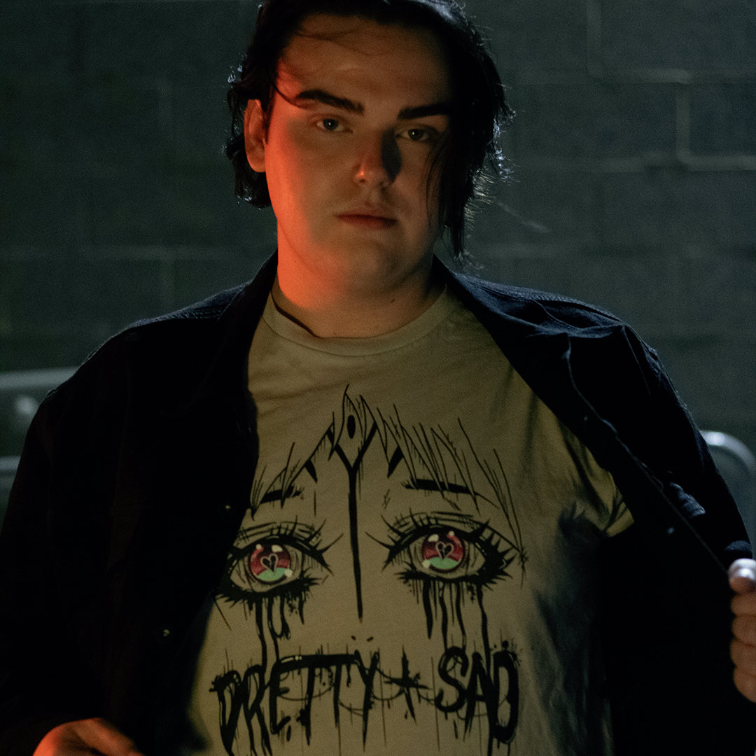 Cream color shirt with a graphic print of stylized eyes and the text 'Pretty & Sad' on the front. Worn by a model in front of a brick wall.