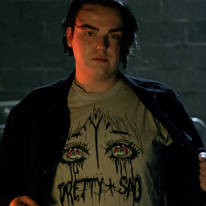 Cream color shirt with a graphic print of stylized eyes and the text 'Pretty & Sad' on the front. Worn by a model in front of a brick wall.