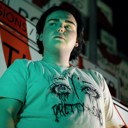 Cream color shirt with a graphic print of stylized eyes and the text 'Pretty & Sad' on the front. Worn by a model in front of street signs.