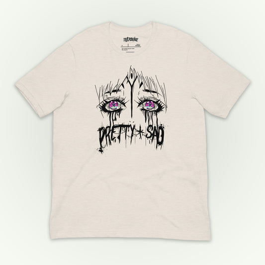 Cream color shirt with a graphic print of stylized eyes and the text 'Pretty & Sad' on the front. flat lay on white background.