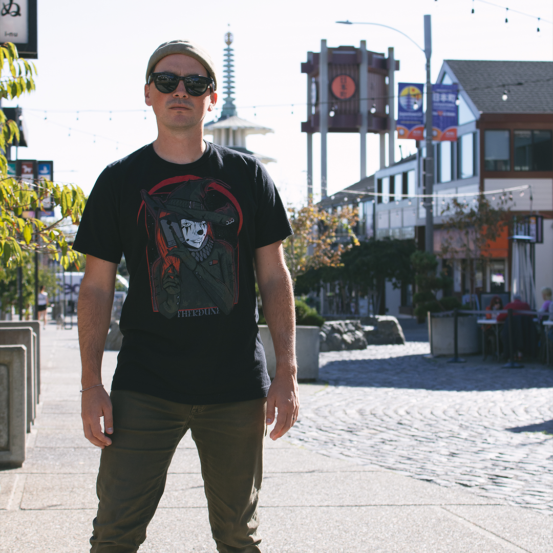 Black shirt with a graphic of a witch clown holding a tarrot card. Worn by a model in an urban environment.