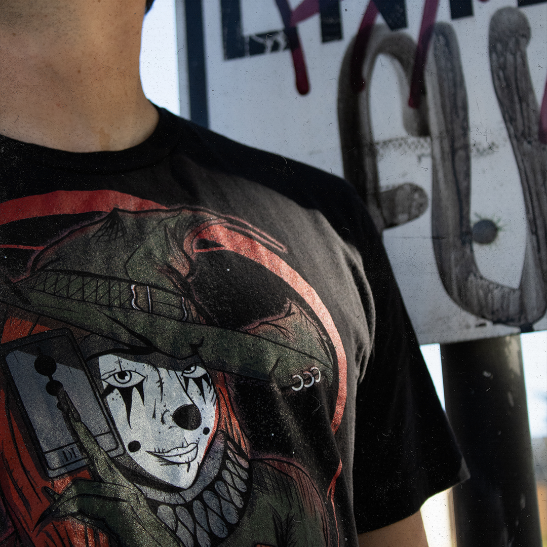 Black shirt with a graphic of a witch clown holding a tarrot card. Worn by a model in front of a street sign covered in graffiti.