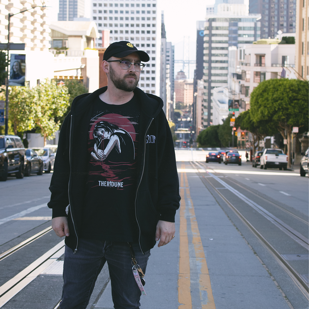 Black shirt with a graphic of a woman crying in knee high water with a red moon in the background. Worn by a model in an urban city.