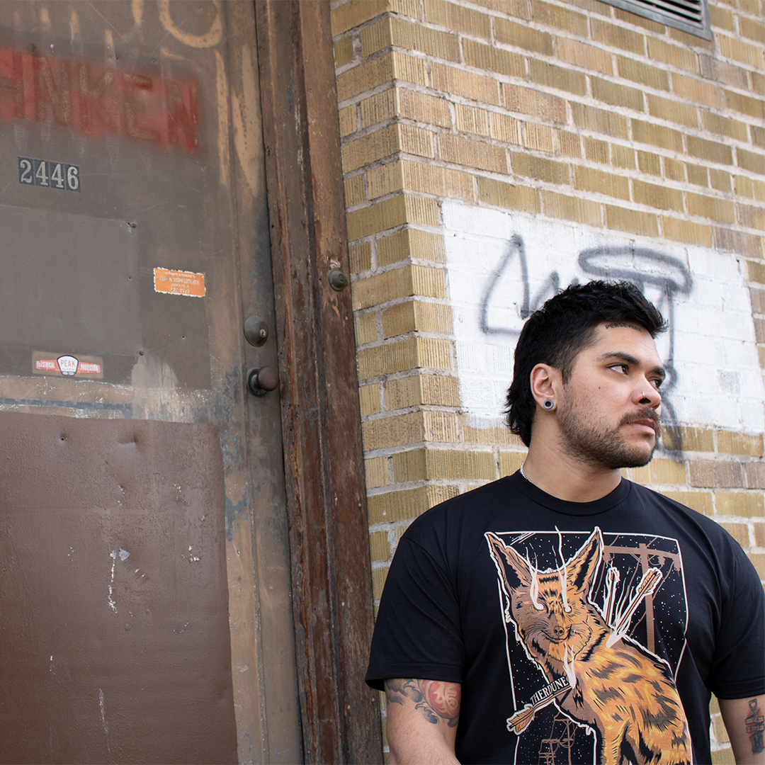 Black shirt with a graphic of a fox impaled with arrows. Worn by a model in front of a brick wall.
