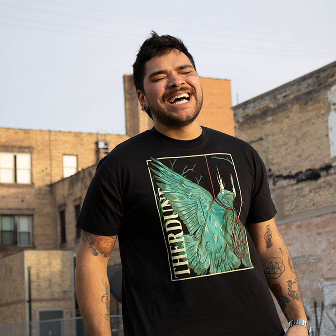 stylized graphic of a green bird, one wing and neck is wrapped up with a red piece of string. "Therdune" logo written on left side vertically. Worn by model in an urban setting.