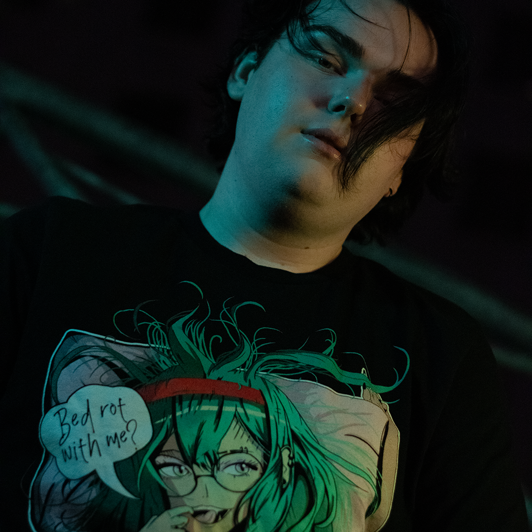 Black shirt with anime style woman, teal hair and bra, with speech bubble saying "Bed Rot With Me?", worn by model, front view.