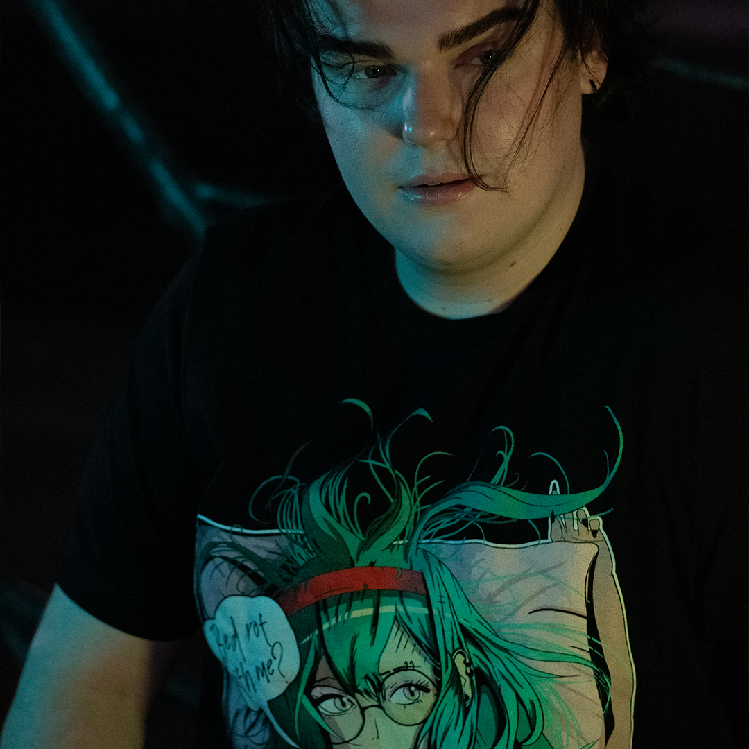 Black shirt with anime style woman, teal hair and bra, with speech bubble saying "Bed Rot With Me?", worn by model, sitting on bench.