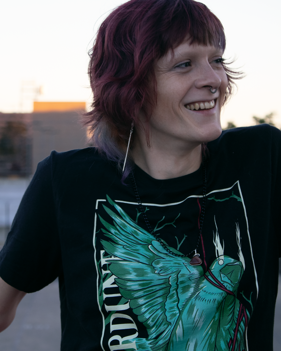 stylized graphic of a green bird, one wing and neck is wrapped up with a red piece of string. "Therdune" logo written on left side vertically. Worn by model outside at dusk.