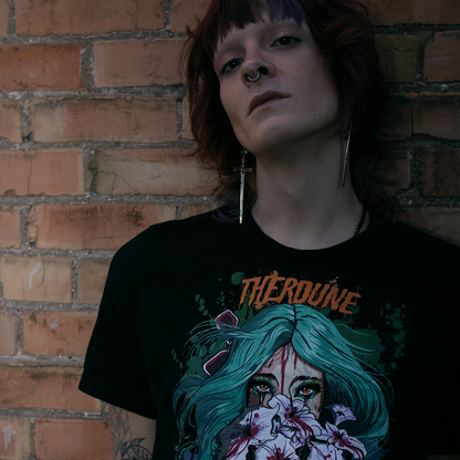 Black shirt with a stylized illustration of a person with teal hair and colorful makeup, worn by a model in front of a brick wall.