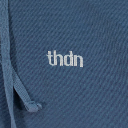 Close-up of a blue jean colored hooded sweatshirt laid flat. A white "thdn" logo is embroidered on the fabric.