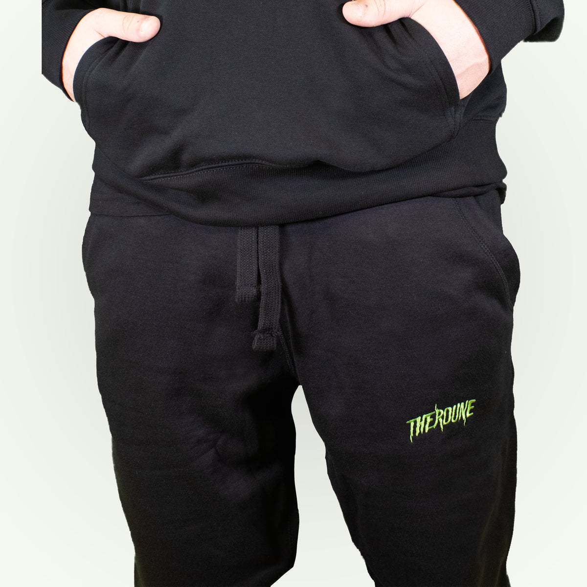 Black sweatpants with a small lime green 'Therdune' embroidered logo on the lower left leg, shown on a model with hands in pockets.