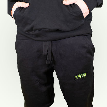 Black sweatpants with a small lime green 'Therdune' embroidered logo on the lower left leg, shown on a model with hands in pockets.